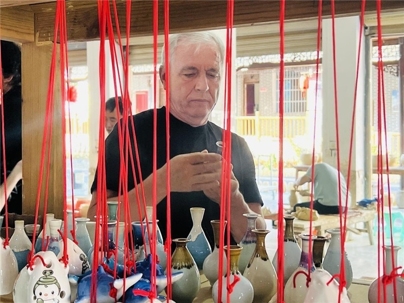 鄉(xiāng)愁如同一根根紅線，將游子與故鄉(xiāng)永遠(yuǎn)地牽連到一起?；逄账囄幕瘎?chuàng)意一條街的母氏陶藝館內(nèi)，紅線吊著陶瓶，思緒游走萬(wàn)里。摩爾多瓦“信息標(biāo)簽”通訊社社長(zhǎng)亞歷山大·塔納斯端詳著館內(nèi)展示的陶藝作品，并挑選出心儀的小擺件帶回摩爾多瓦，也帶走花茂村的“鄉(xiāng)愁”。（張亞欣攝）